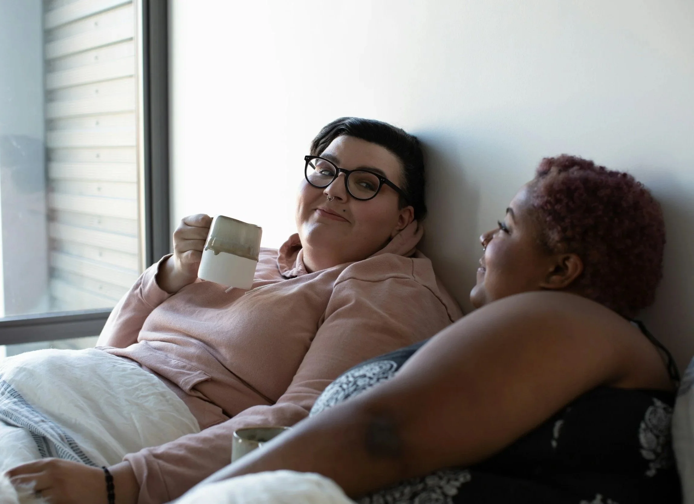 Queeres, lesbisches, trans, nicht-binäres Paar kuschelnd gemeinsam im Bett mit Kaffee | queersensible Paarberatung | Paartherapie | systemische Beratung | psychologische Beratung | Annu Jona Winkler