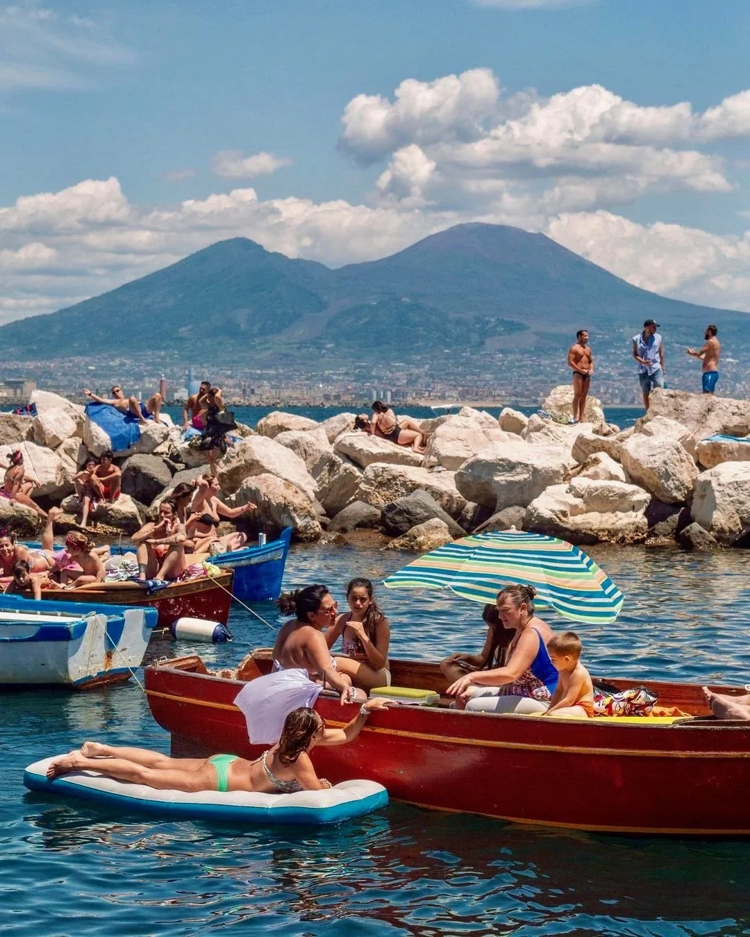 People enjoying a sunny day by the lake, some on boats, others sitting and lying on rocks and on a float, with mountains in the background and partly cloudy sky.