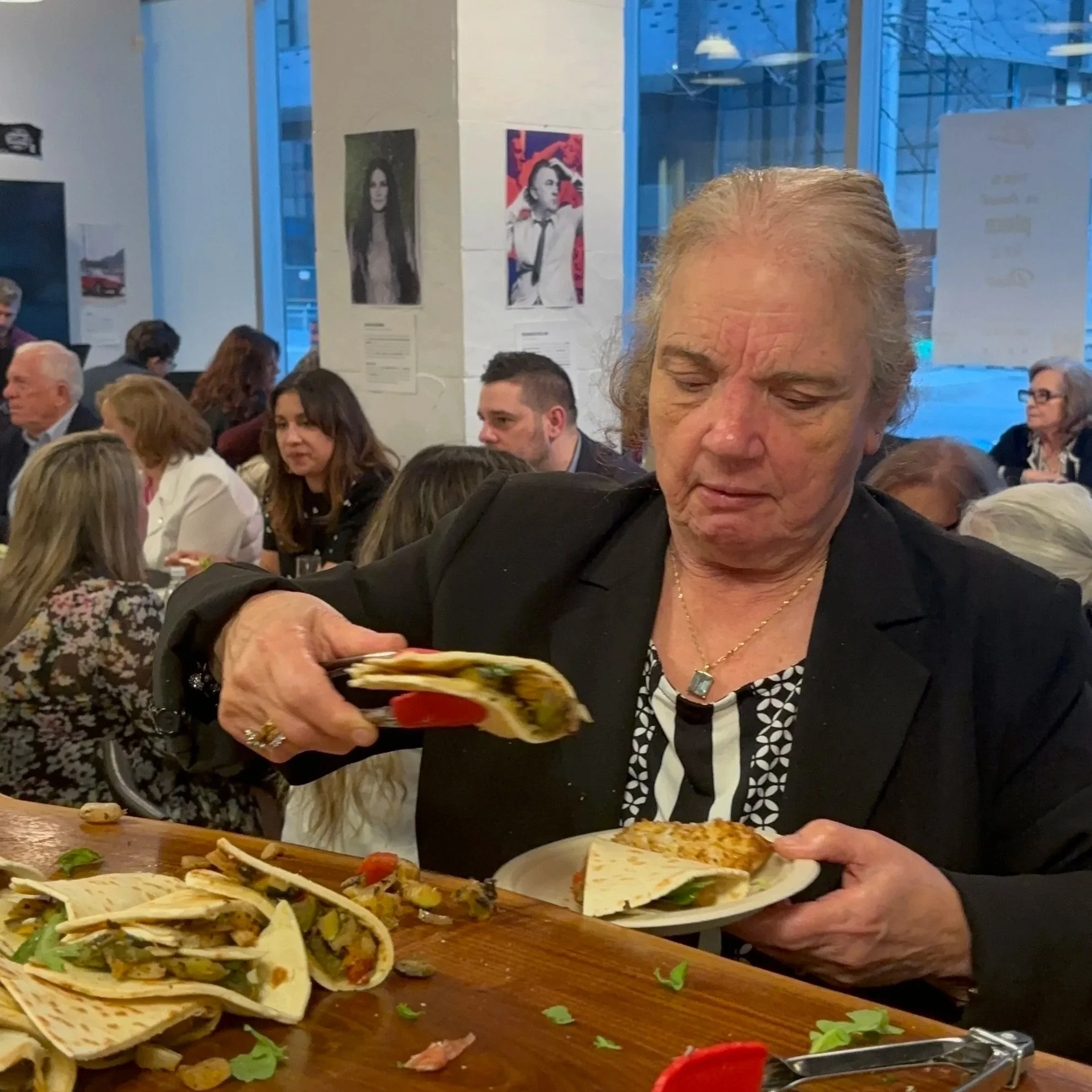People sitting at a table in a restaurant, with a tray of tacos and dishes in the foreground. A man with gray hair is in focus, looking down as he holds a plate. Other diners are visible in the background, and restaurant decor includes wall art and l
