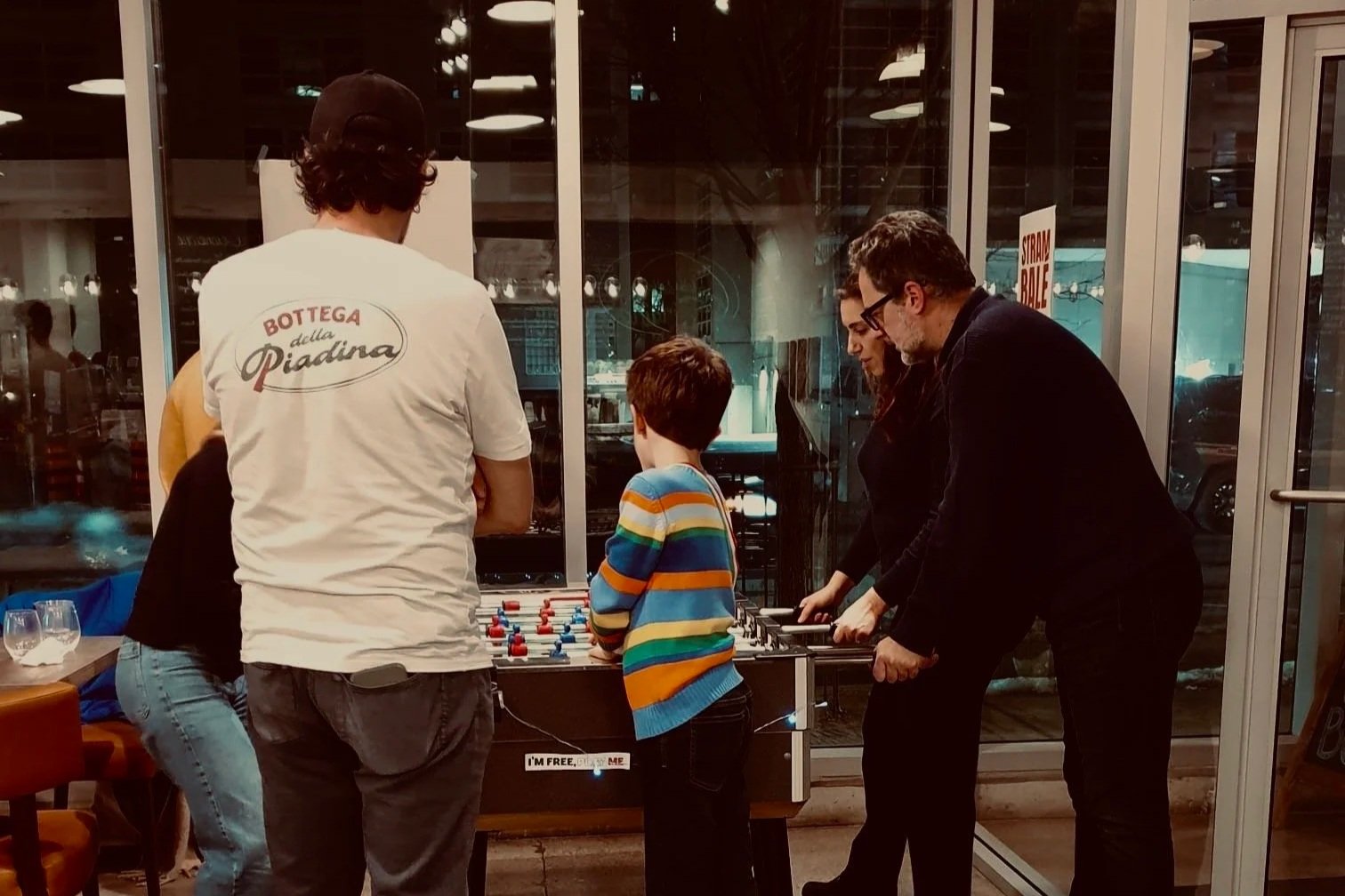 Group of four people playing foosball at a restaurant or cafe with large glass windows in the background.
