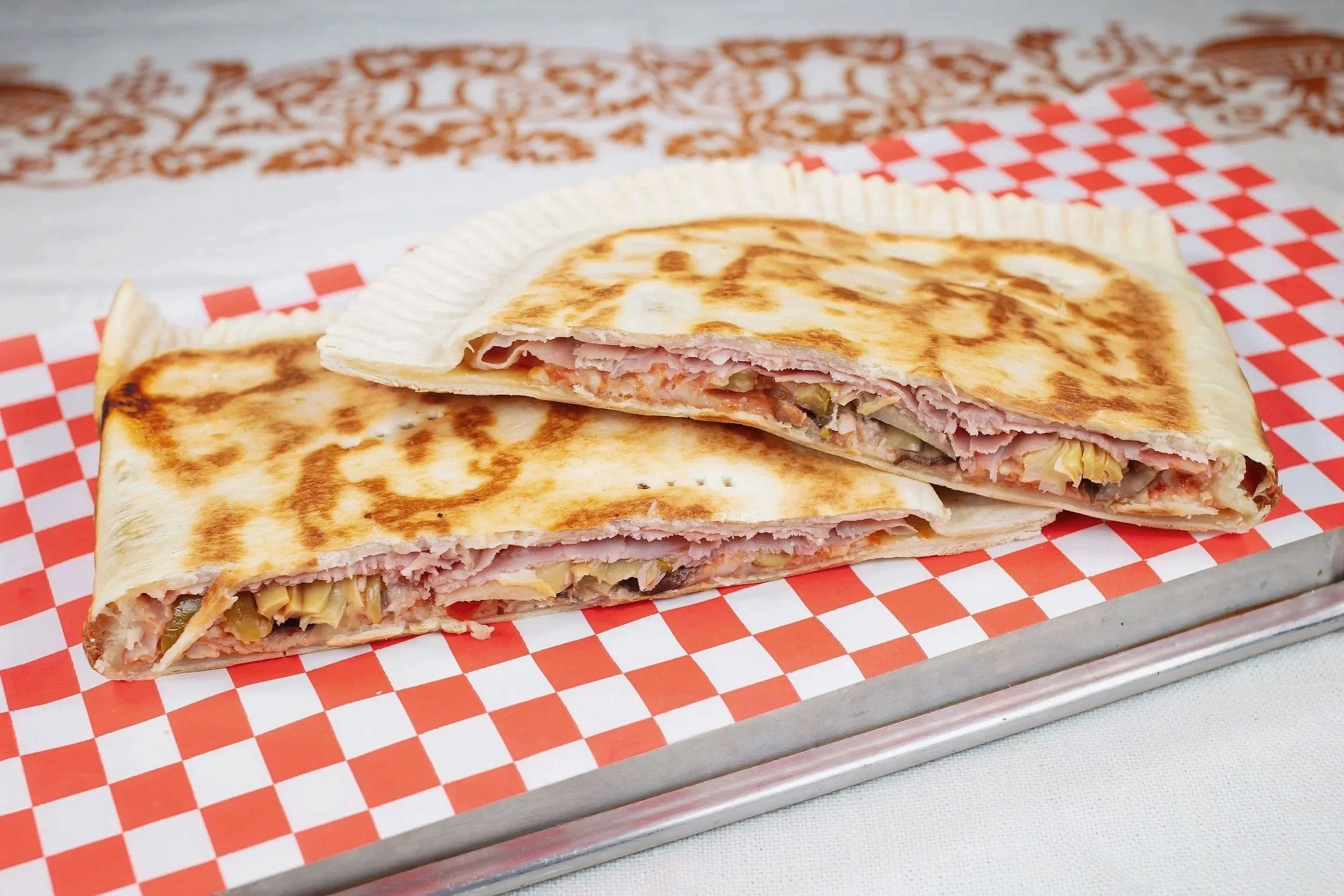 Close-up of a grilled wrap sandwich with ham, cheese, and vegetables, cut in half, placed on red and white checkered paper on a white surface.