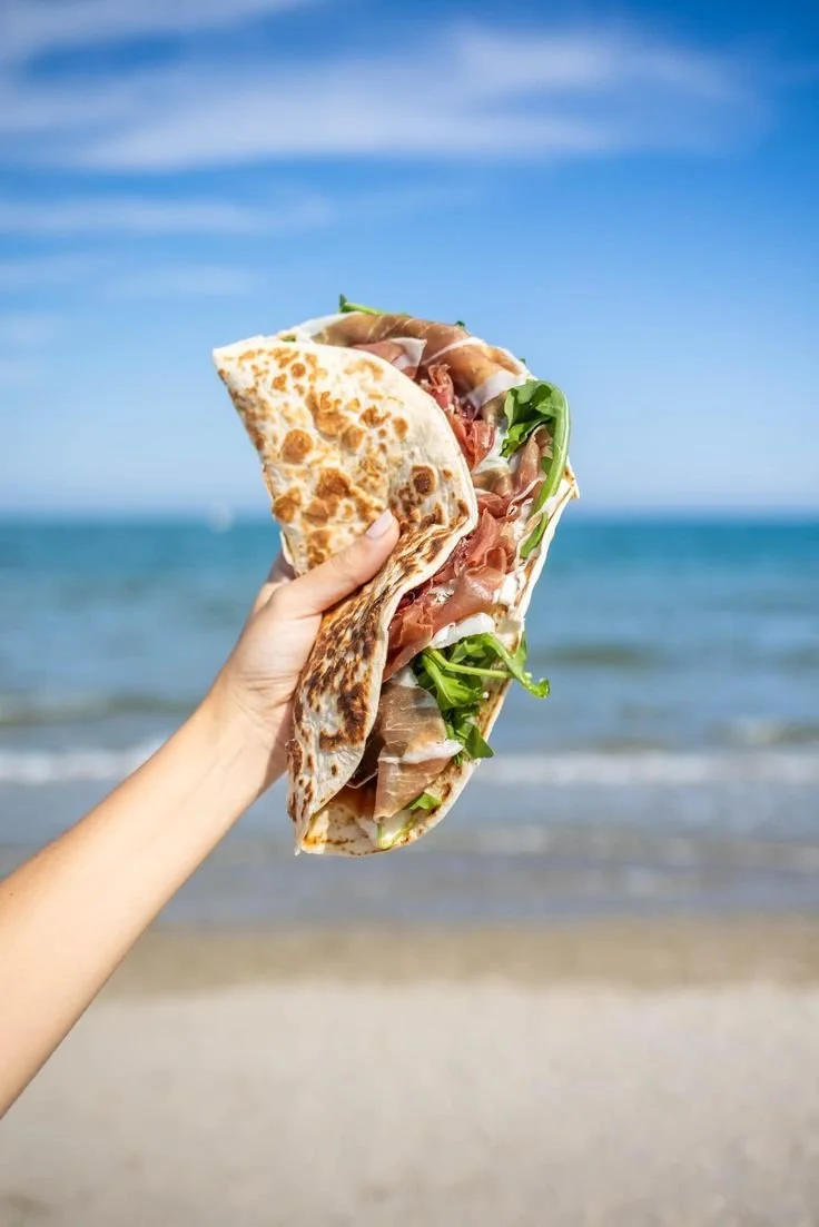 A hand holding a piadina filled with prosciutto and arugula by the seaside