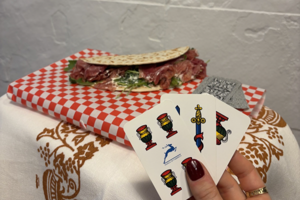 Hand holding four playing cards with goblets and swords, in front of a wrapped sandwich with meat and greens, on a table with patterned tablecloth and a gray wall background.