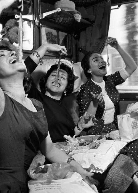 Three women sitting inside a bus, holding french fries to their noses and smiling, with food and bags in front of them.