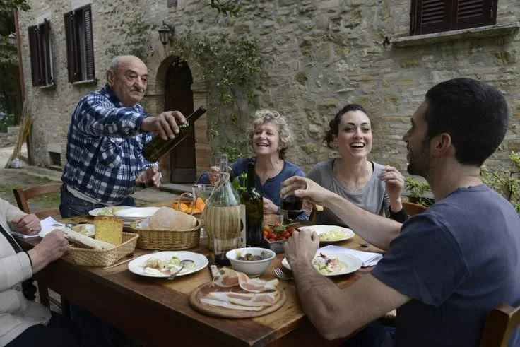Group of five people enjoying a meal outdoors at a wooden table, with food and drinks, in a rustic setting with stone walls.
