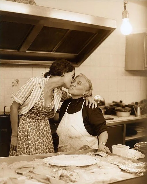 Two women in a kitchen, one giving the other a kiss on the cheek, surrounded by cooking utensils and food.
