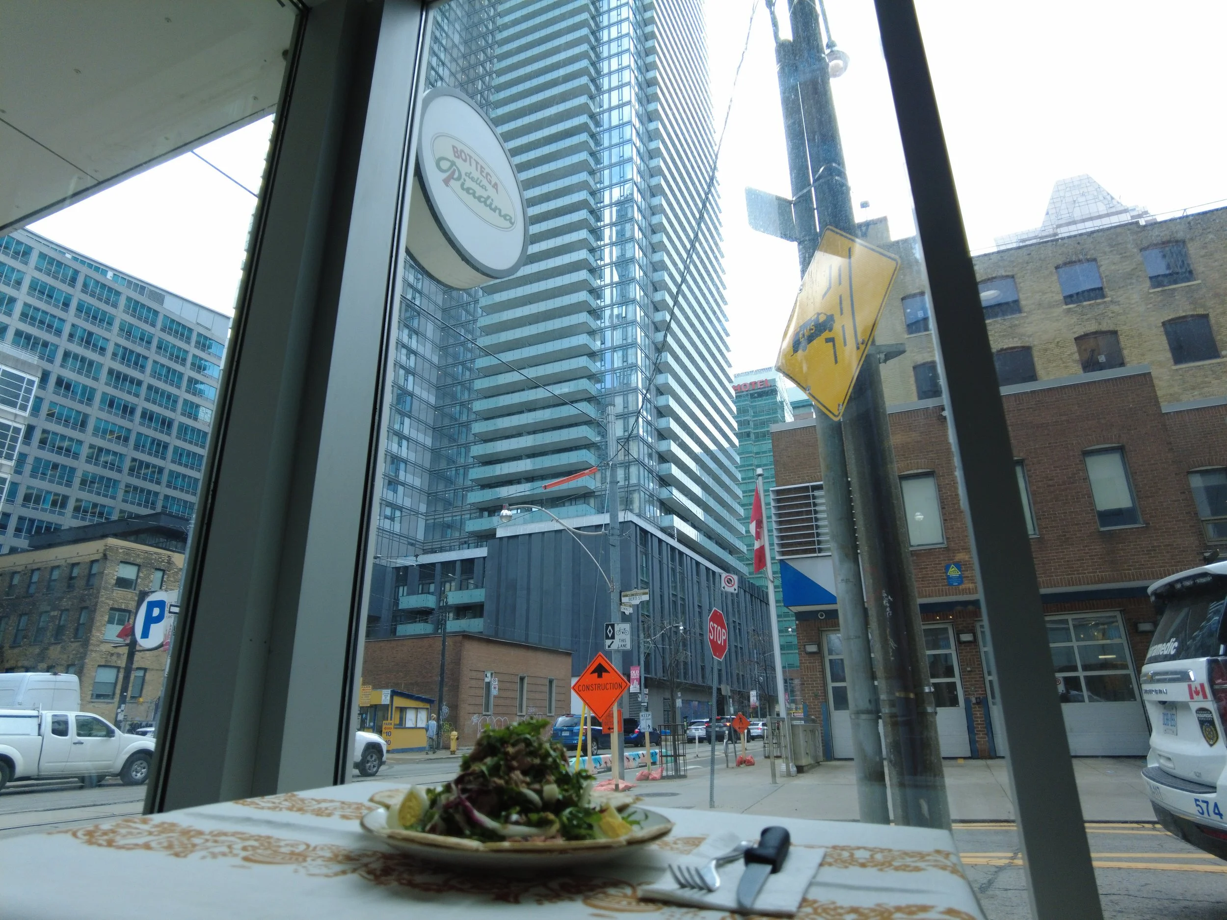 View of tall modern buildings seen from inside a restaurant or cafe. There is a plate of food on the table, and various street signs and construction signs are visible outside.
