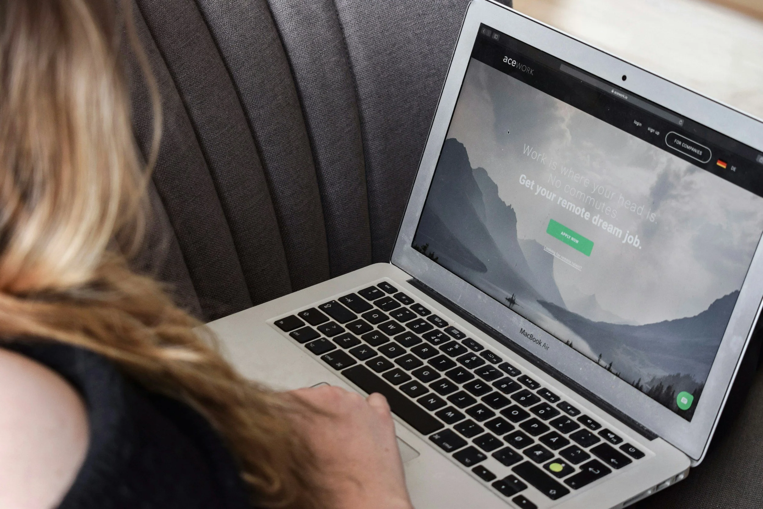 Persoon die op een MacBook Air laptopscherm kijkt, waarop een website voor remote werken te zien is, met bergen op de achtergrond en een groene knop 'Apply Now'.