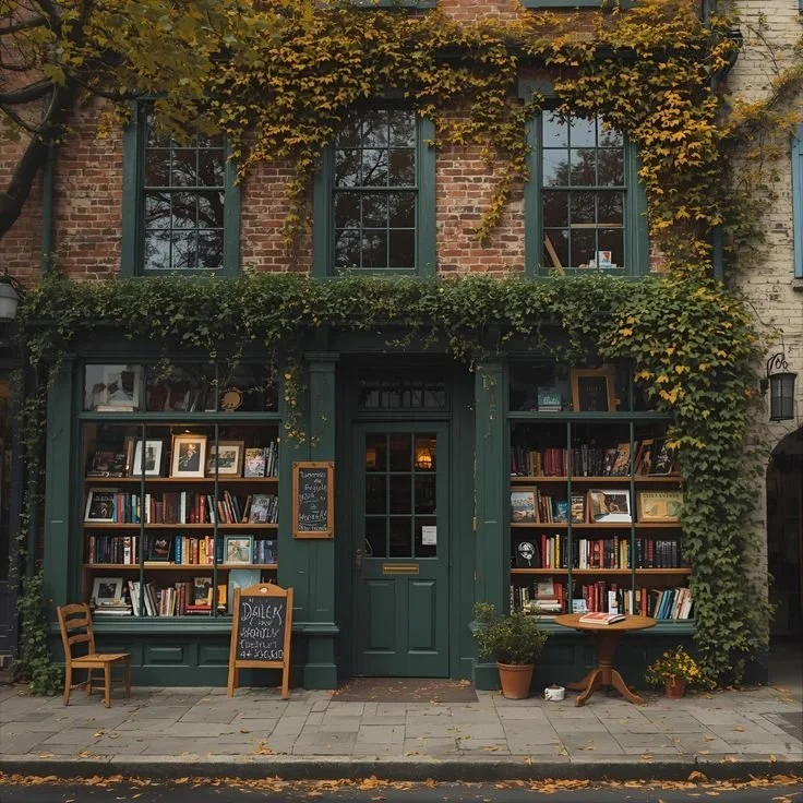 A cozy bookstore with a green facade and large windows, surrounded by autumn leaves, with books displayed on open shelves and extras outside.