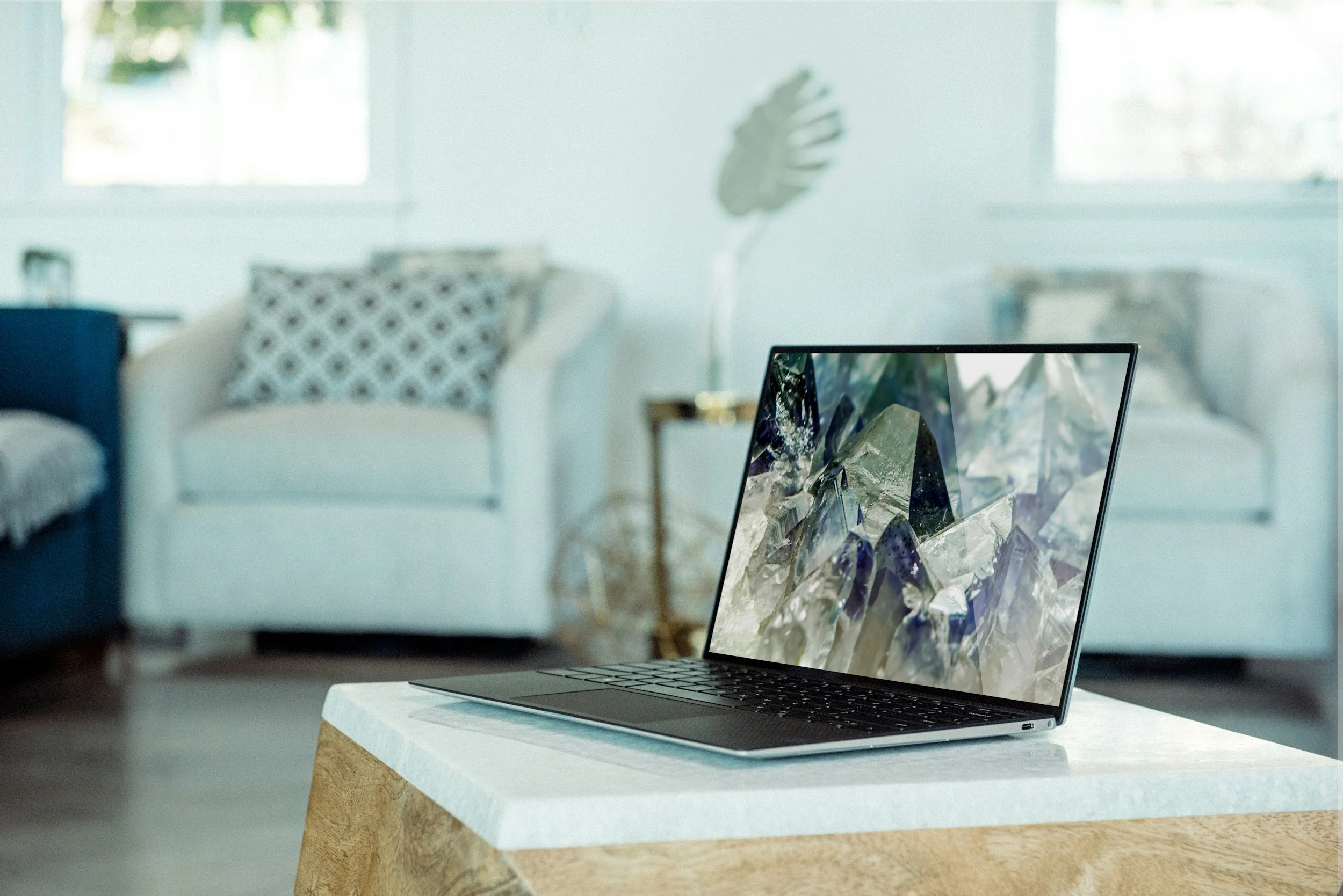 Slim laptop open on a white and wood coffee table in a bright living room with sofas and large windows