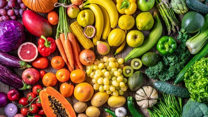 Assorted fresh fruits and vegetables on a wooden surface, including bananas, apples, grapes, carrots, peppers, broccoli, zucchini, and more.