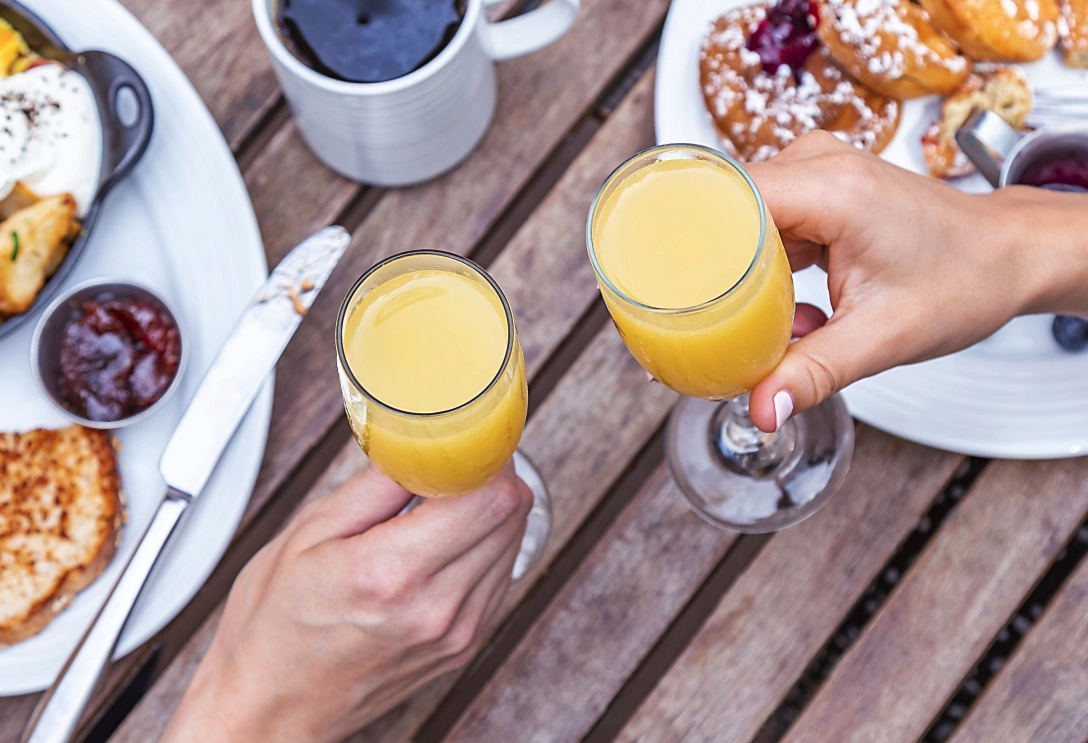 Two people clinking glasses of orange juice at an outdoor breakfast table with plates of food and coffee.