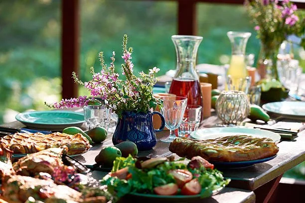 A table set for a meal with flowers in a blue pitcher, pitchers of juice, plates, glasses, and dishes of salad and pie outdoors with greenery in the background.