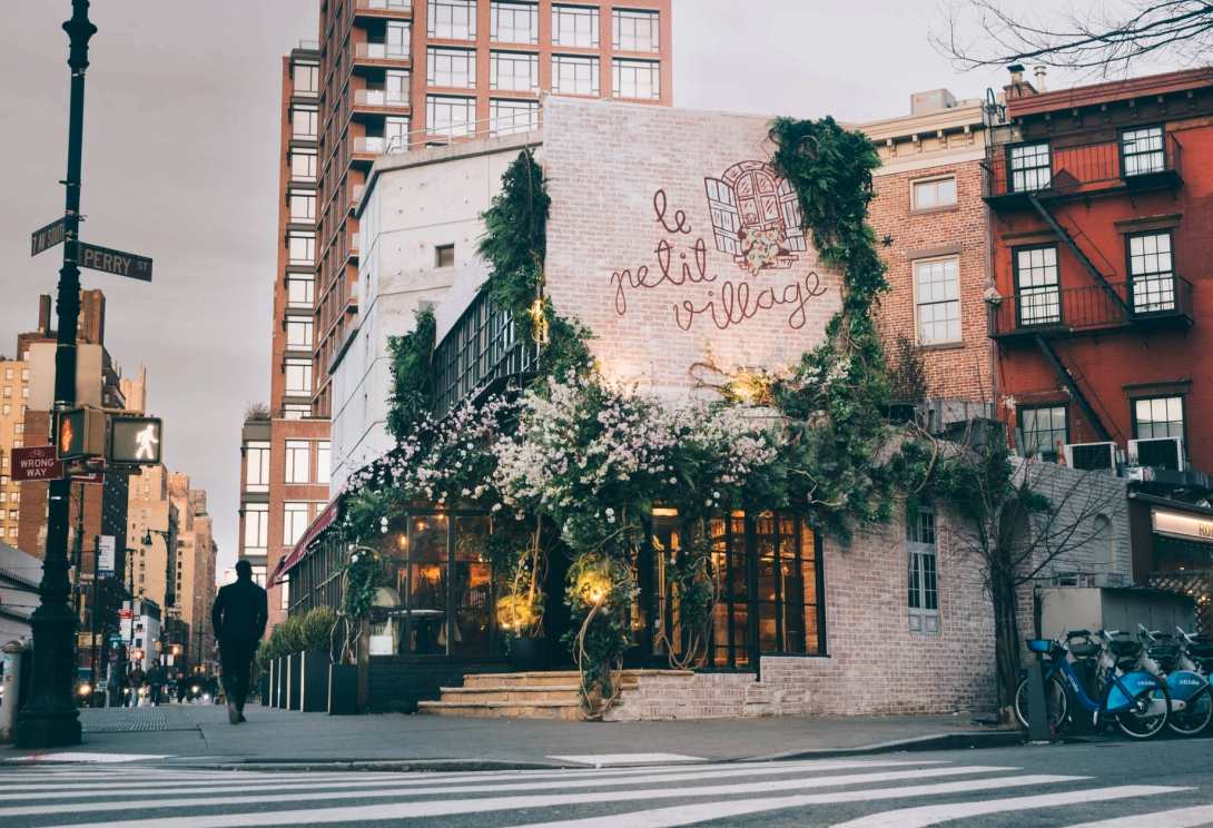 Street scene featuring a building with a sign that reads 'Le Petit Village' and decorative greenery, with pedestrians and bicycles in the vicinity.