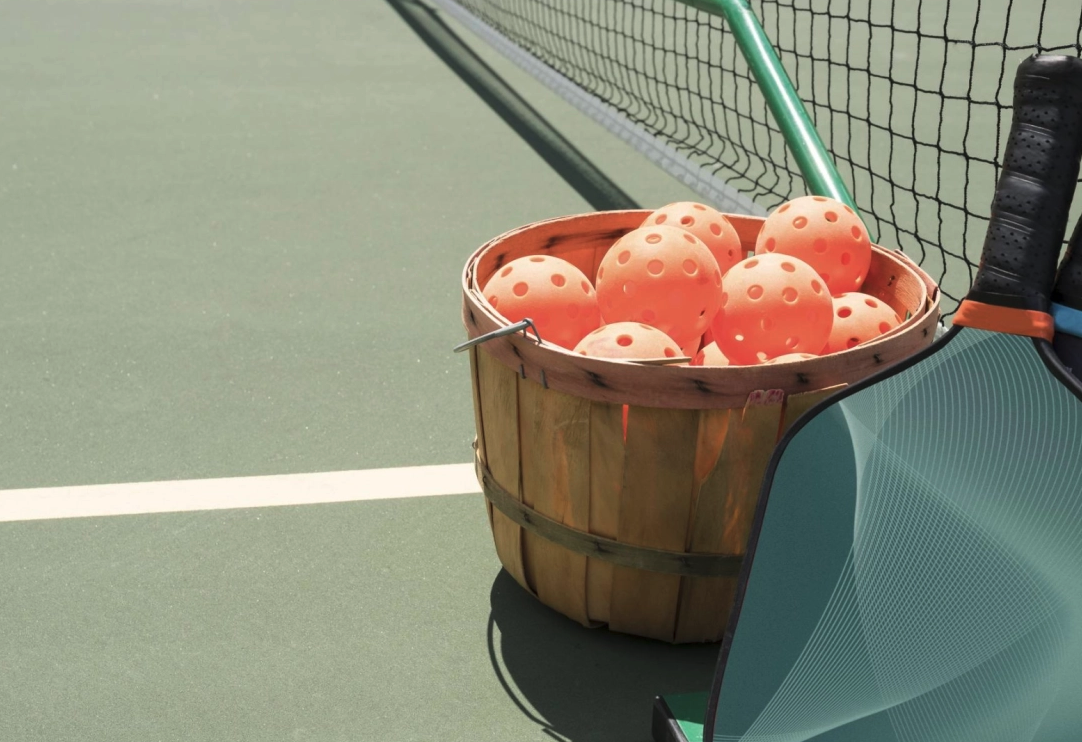 A wooden basket filled with pink rubber tennis balls on a tennis court, with a tennis racket leaning against the basket.