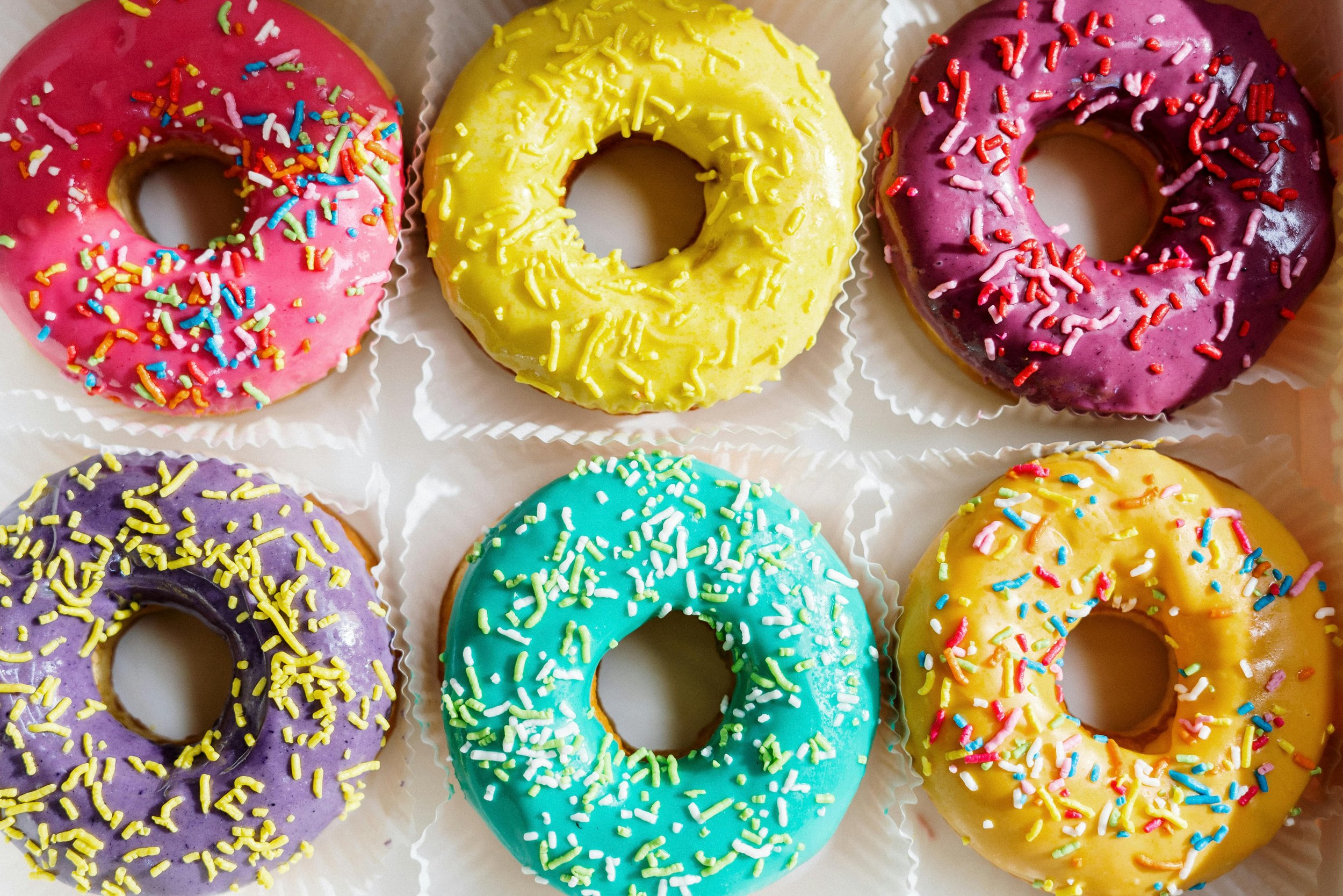 Six colorful donuts with sprinkles on a white tray, arranged in two rows of three. The donuts are pink, yellow, purple, blue, purple, and yellow with various sprinkles.