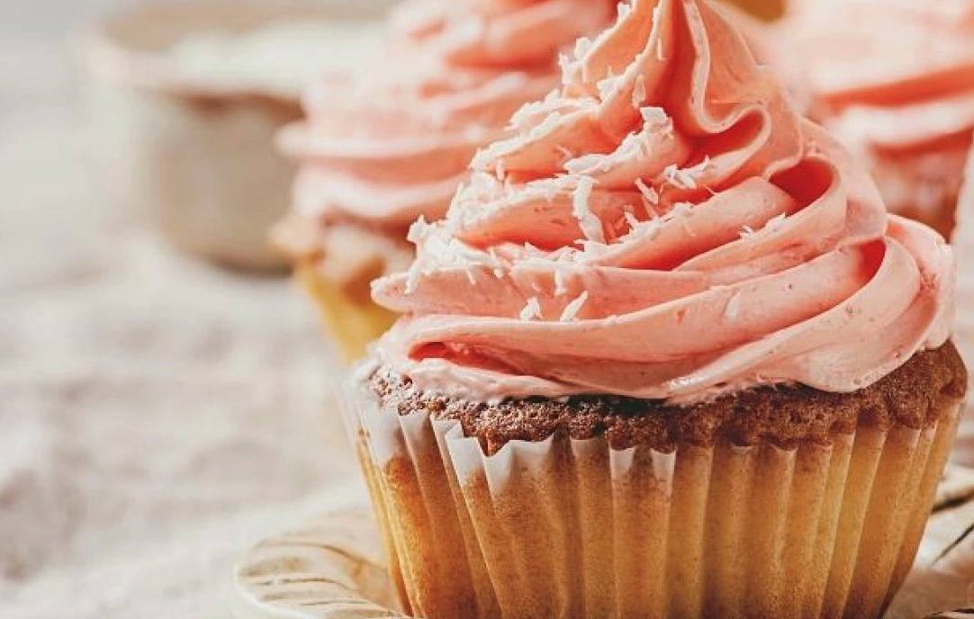 Close-up of a cupcake with pink frosting and white shavings, placed on a rustic surface.