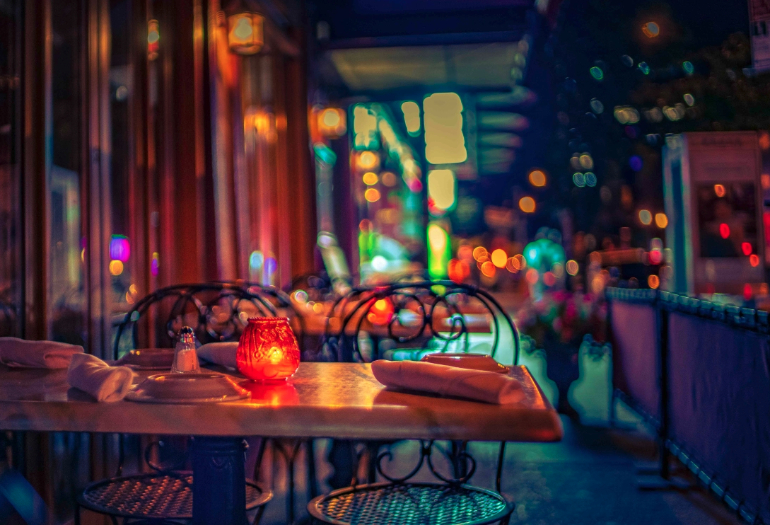 Empty outdoor restaurant table illuminated by a red candle with a napkin, set with plates and utensils, at night with blurred colorful city lights in the background.