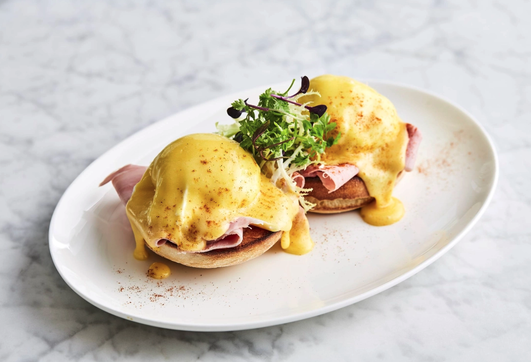 Eggs Benedict with ham and microgreens on a white plate on a marble surface.