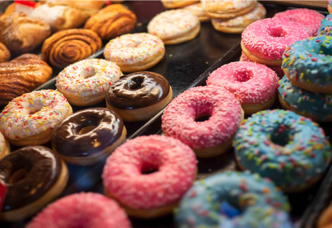 Assorted donuts on display, including glazed, chocolate, strawberry with sprinkles, and colorful frosted varieties.
