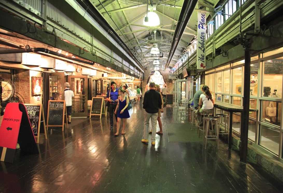 Indoor marketplace or food hall with people walking and sitting at tables, illuminated by hanging lights, with storefronts and signage along the sides.