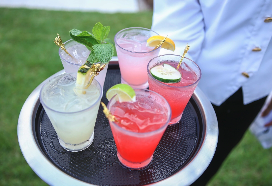 A tray of five colorful cocktails with garnishes, including lemon slices, cucumber slices, mint, and decorative stir sticks, held by a person in a white shirt outdoors.