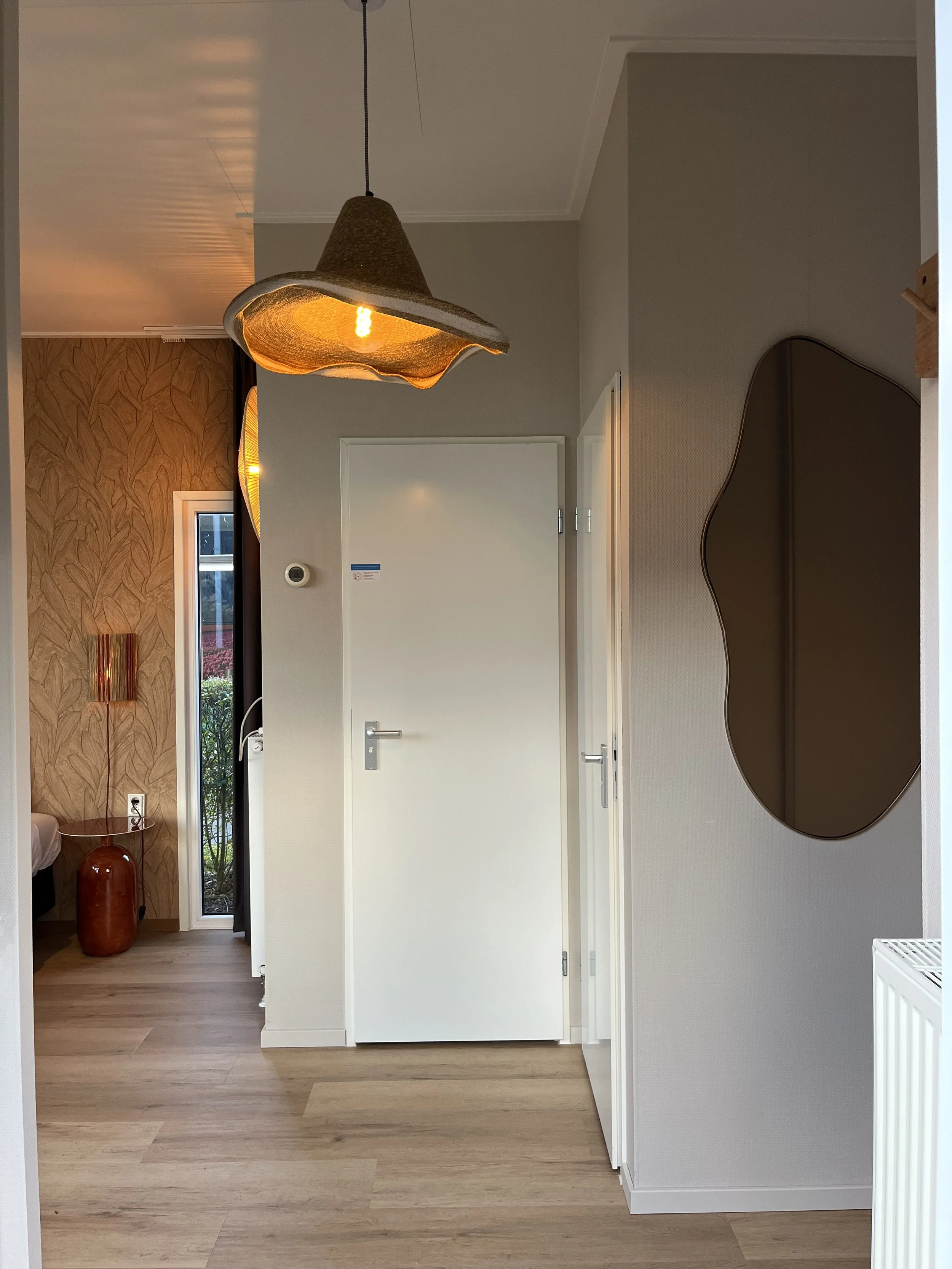 Een woonkamer met een grote gemakkelijke bank, decoratieve muurwandobjecten, een grote plant in een pot, en grote ramen met zwart jaloezieën die zonlicht en schaduw binnenlaten.