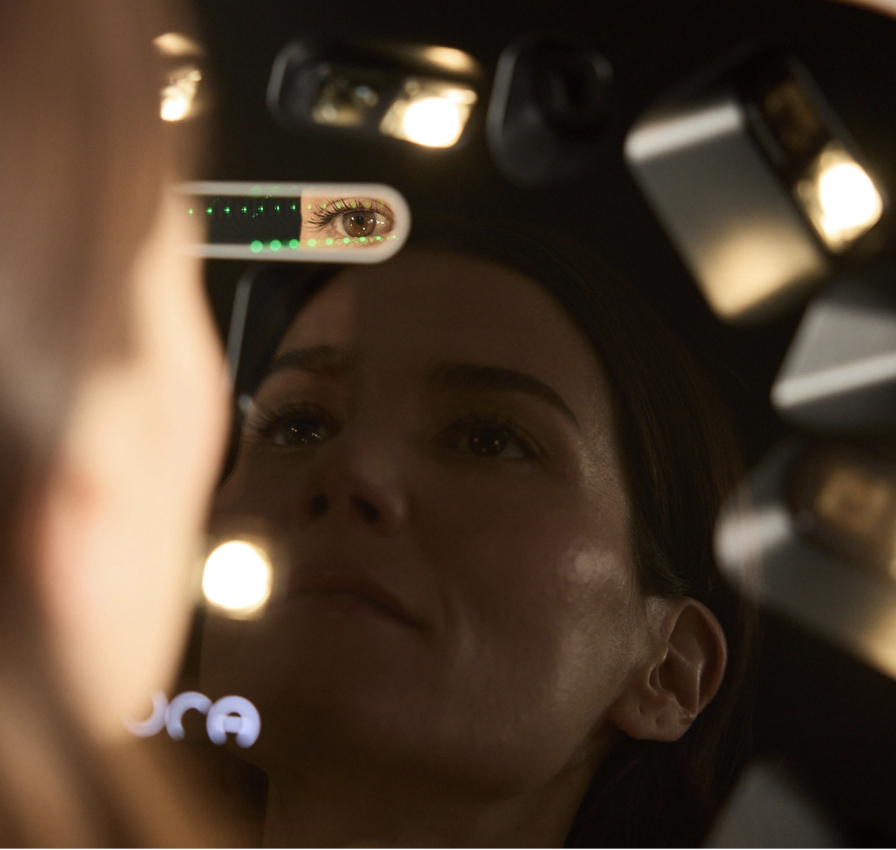 A woman looks into a mirror, with her eye and part of her face reflected in the mirror, surrounded by bright lights.