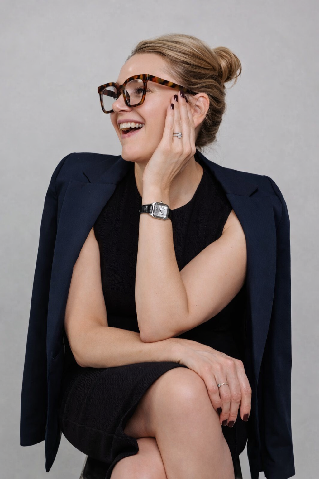 A woman with blonde hair styled in a bun, wearing large tortoiseshell glasses, a black sleeveless dress, and a navy blazer draped over her shoulders. She is smiling, touching her face with her left hand, showing a ring, and has a wristwatch on her left wrist. The background is plain gray.