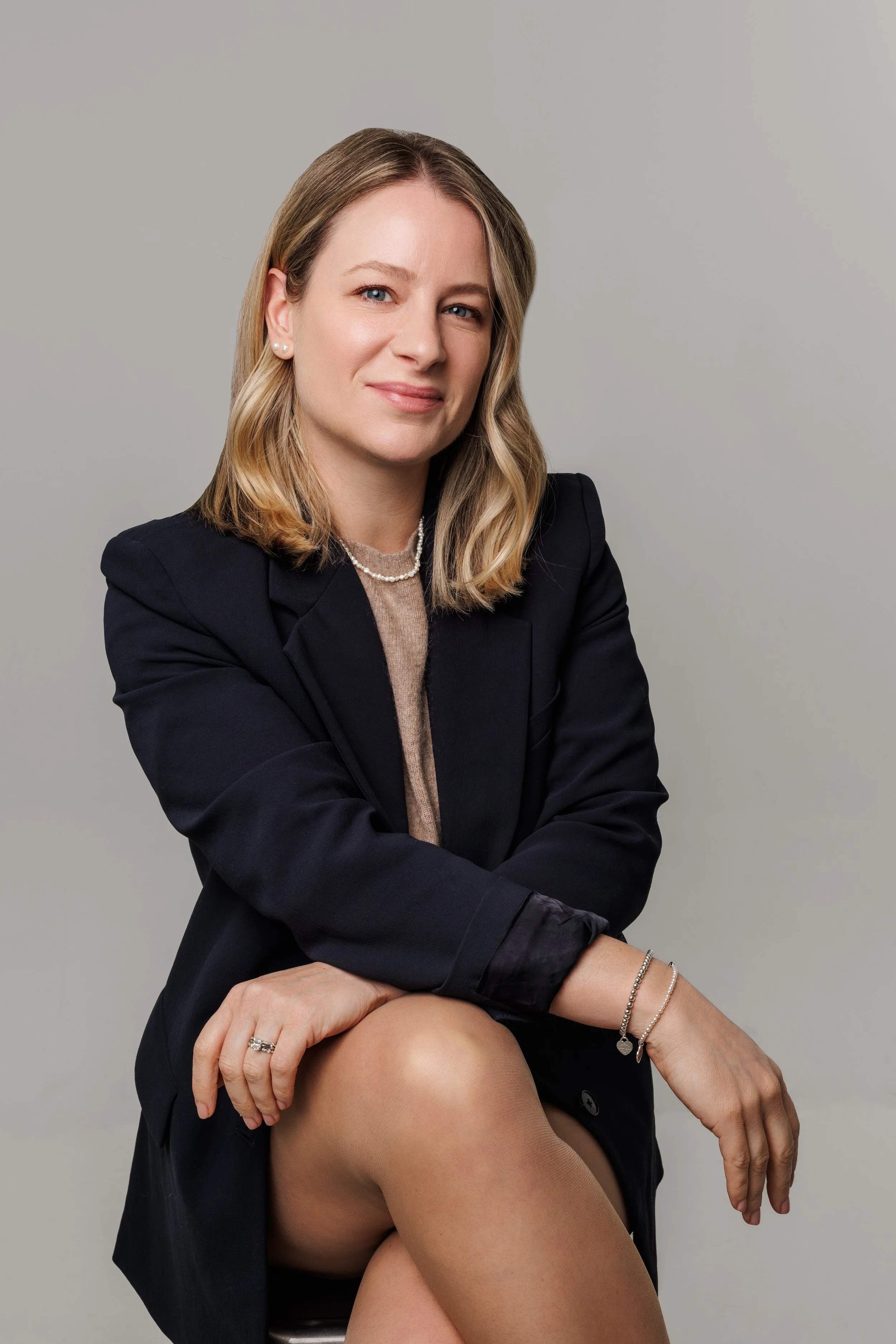 A woman with shoulder-length blonde hair smiling and looking at the camera, sitting with legs crossed, wearing a black blazer, a beige top, jewelry, and a neutral background.