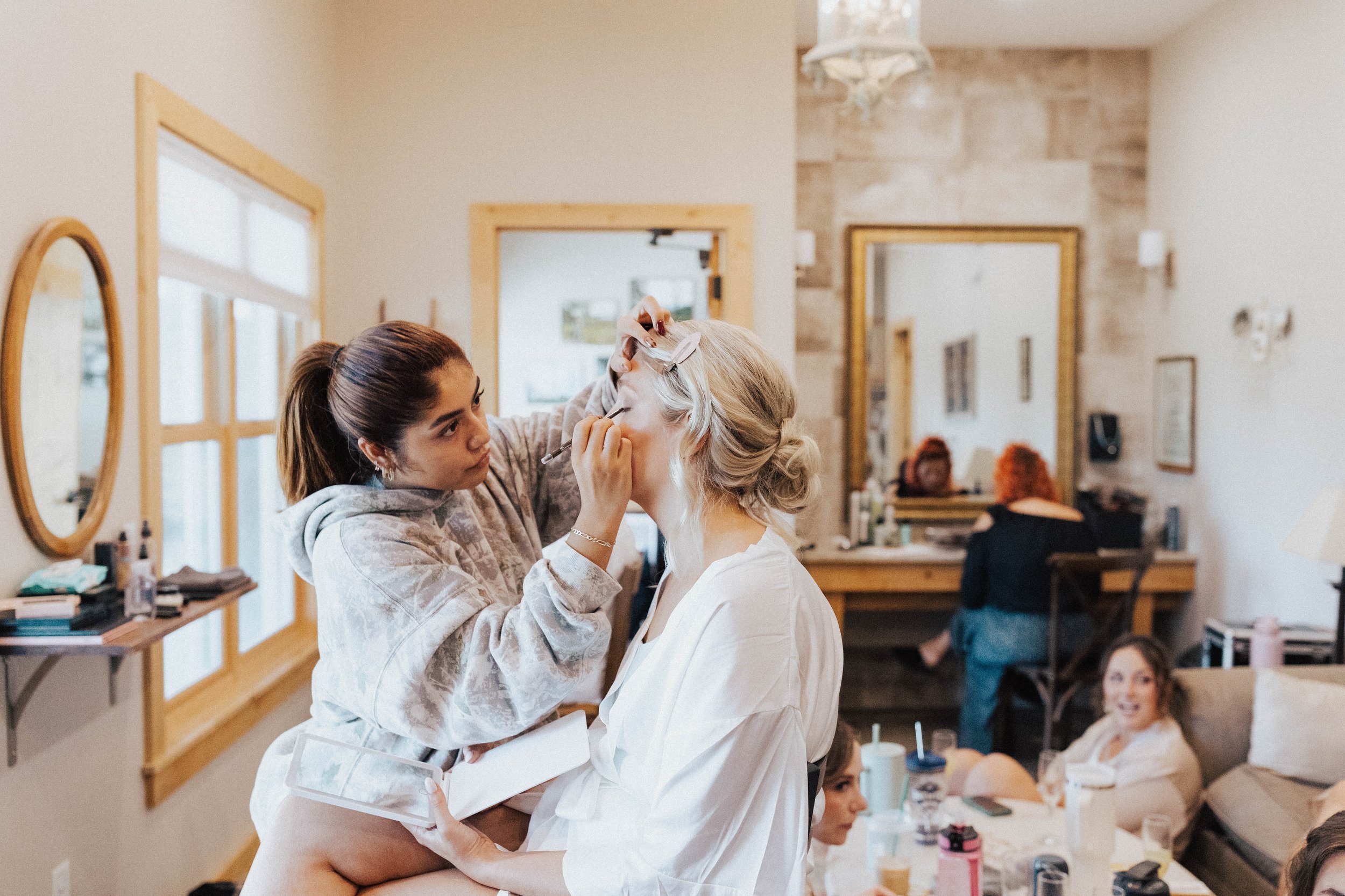 Makeup artist applying makeup to a woman seated in a cozy room with a mirror, while others relax on a sofa and someone works at a desk in the background – bridal prep at One Salon & Bridal Co in St. Charles, IL