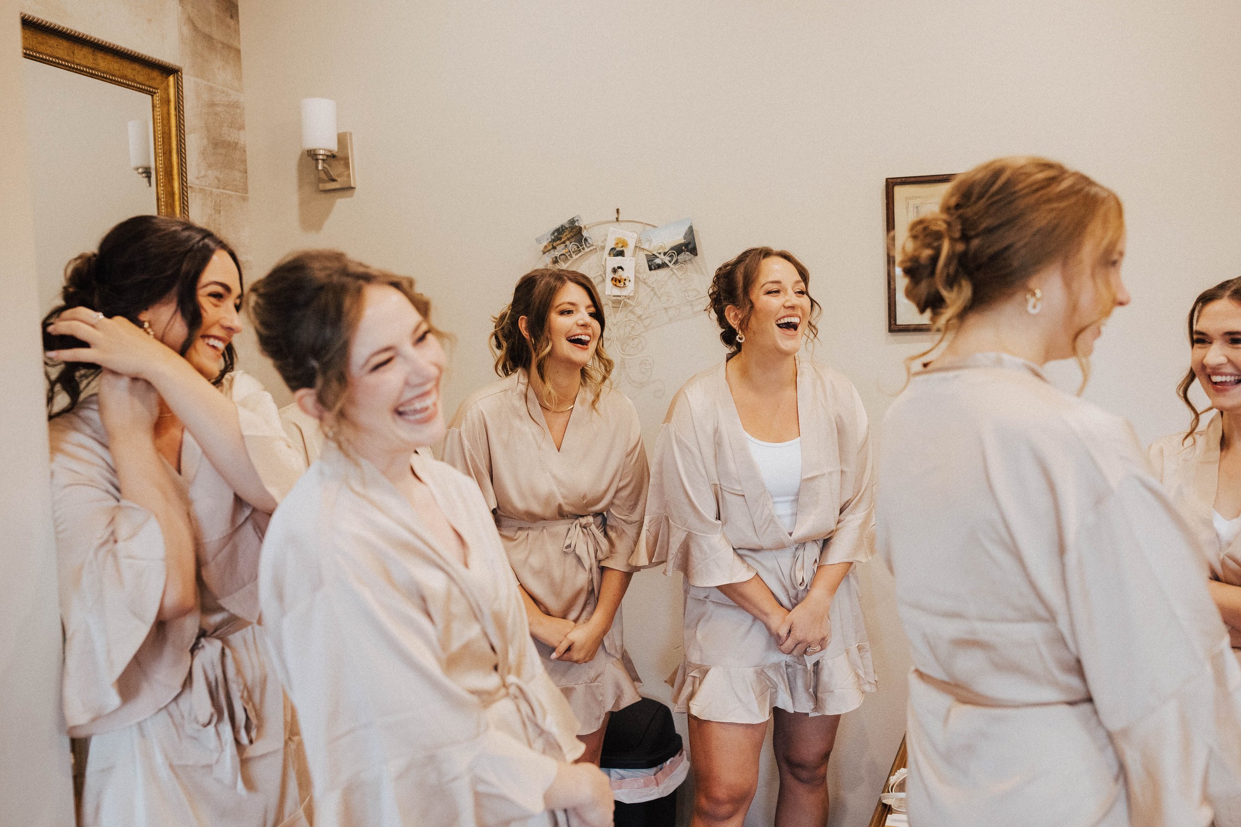 Six women in matching beige robes laughing and smiling while getting ready for a special event – bridal party prep at One Salon & Bridal Co in St. Charles, IL