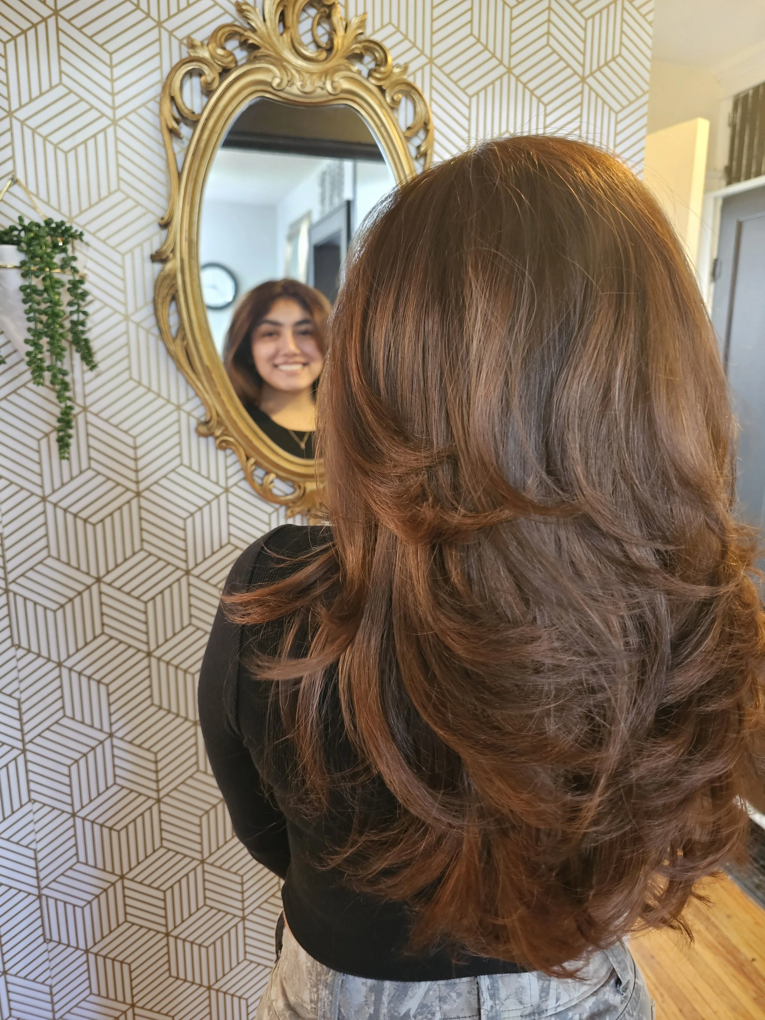 A woman with long, layered, brown hair is looking in a mirror and smiling, with her reflection showing her face– styled at One Salon & Bridal Co in St. Charles, IL