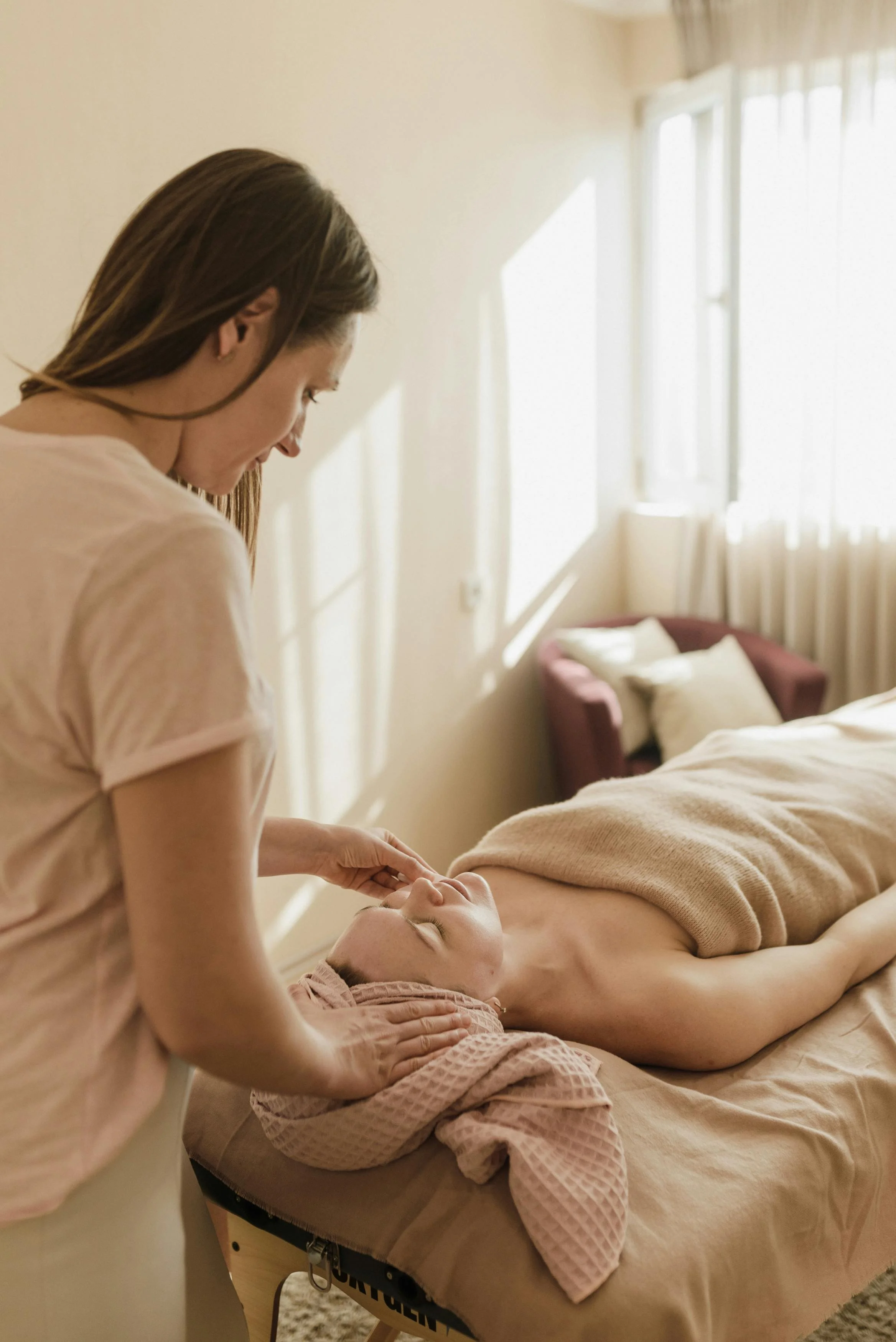 Esthetician performing a facial massage on a client lying on a massage table in a bright, sunlit room at One Salon & Bridal Co in St. Charles, IL