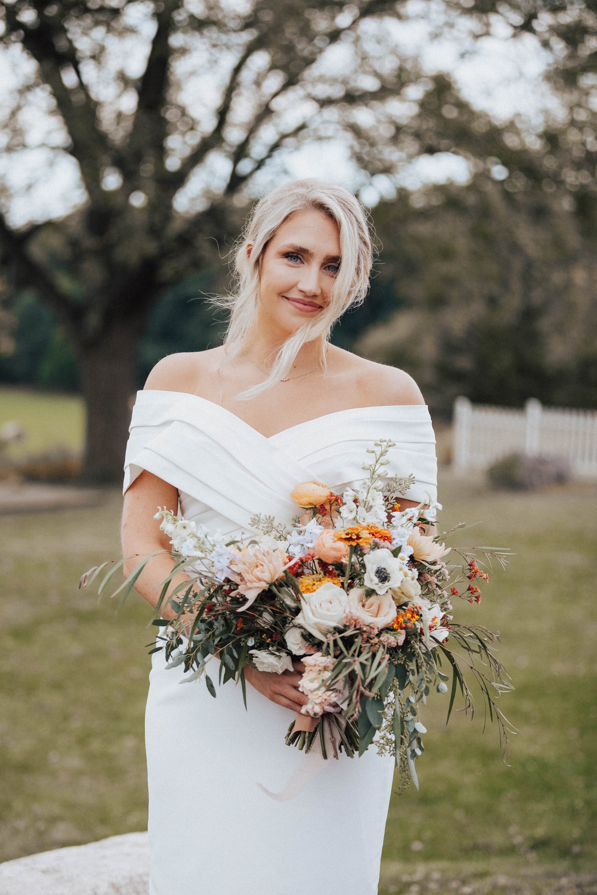 Bride in a white off-shoulder wedding dress holding a large bouquet and smiling outdoors with trees and a fence in the background – styled by One Salon & Bridal Co in St. Charles, IL