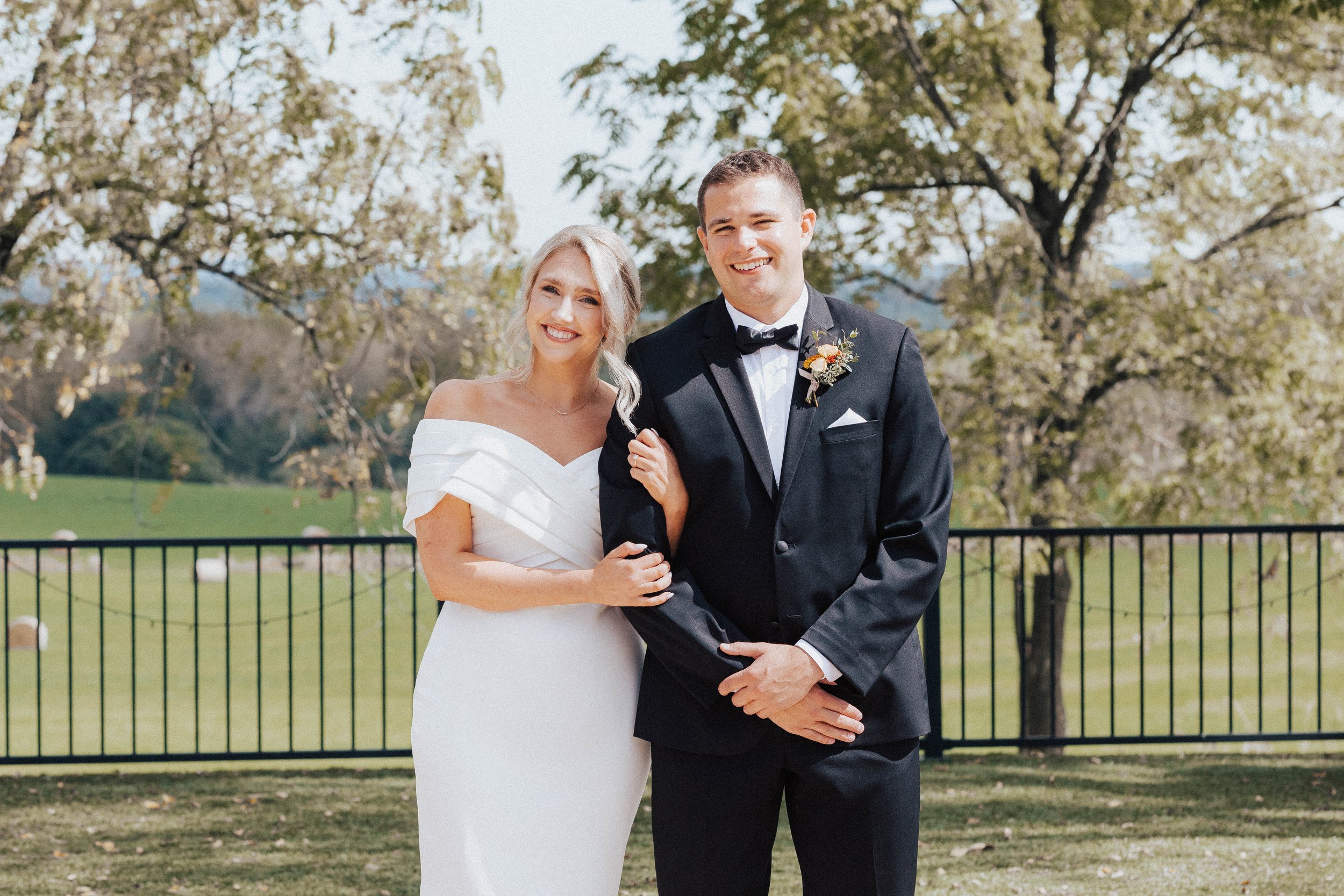 Bride and groom smiling outdoors on wedding day, standing in front of a black fence with trees and green landscape in the background--styled by One Salon & Bridal Co in St. Charles, IL