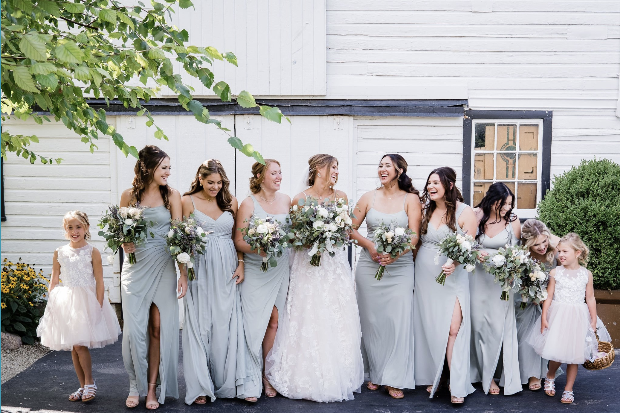 Group of women and girls dressed in light gray and white wedding attire, standing outdoors and laughing together, holding bouquets of flowers -- styled by One Salon & Bridal Co in St. Charles, IL