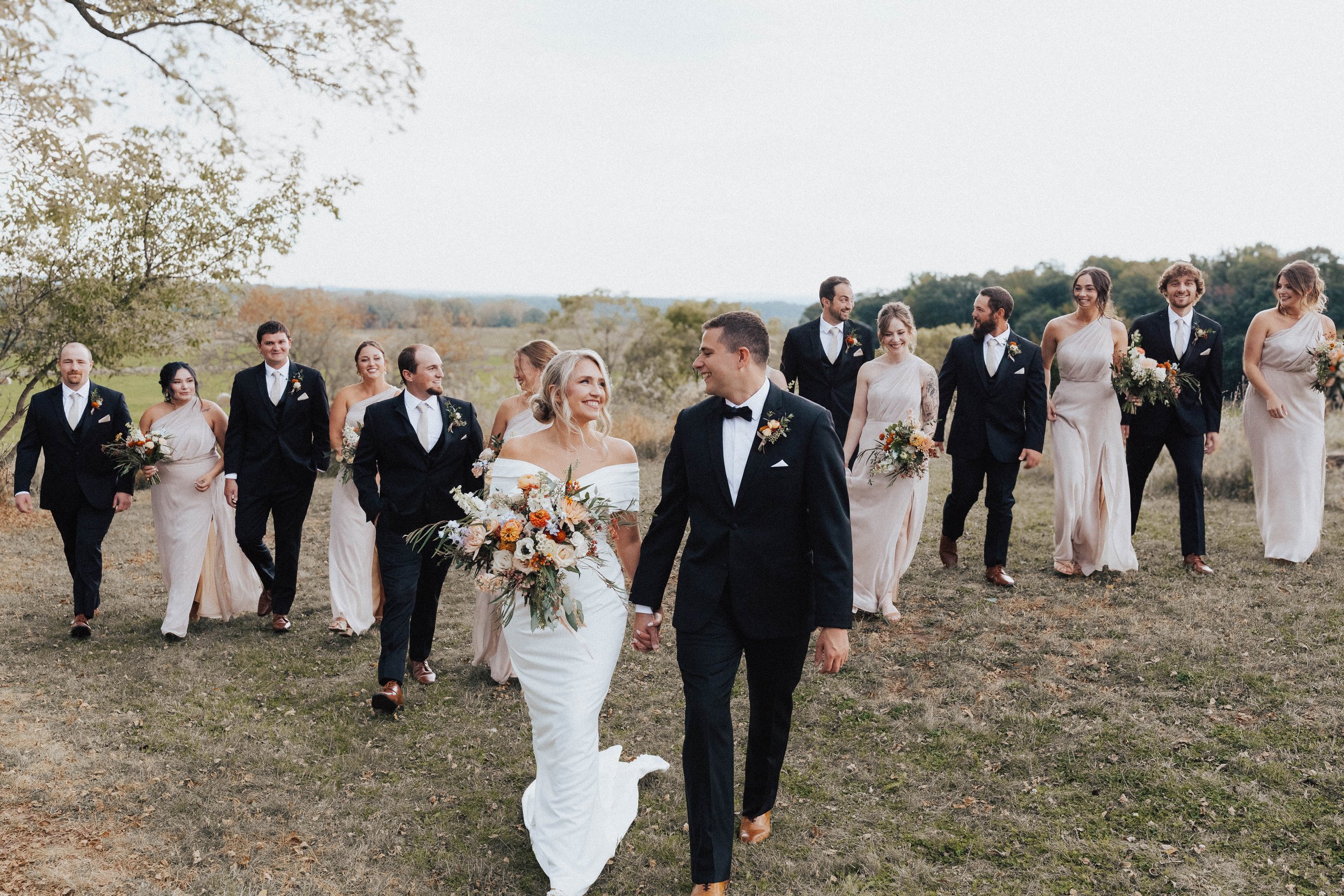 A wedding party walking outdoors on a grassy field, with the bride and groom in front holding hands, everyone dressed in formal attire, with the bride holding a large bouquet of flowers--styled by One Salon & Bridal Co in St. Charles, IL