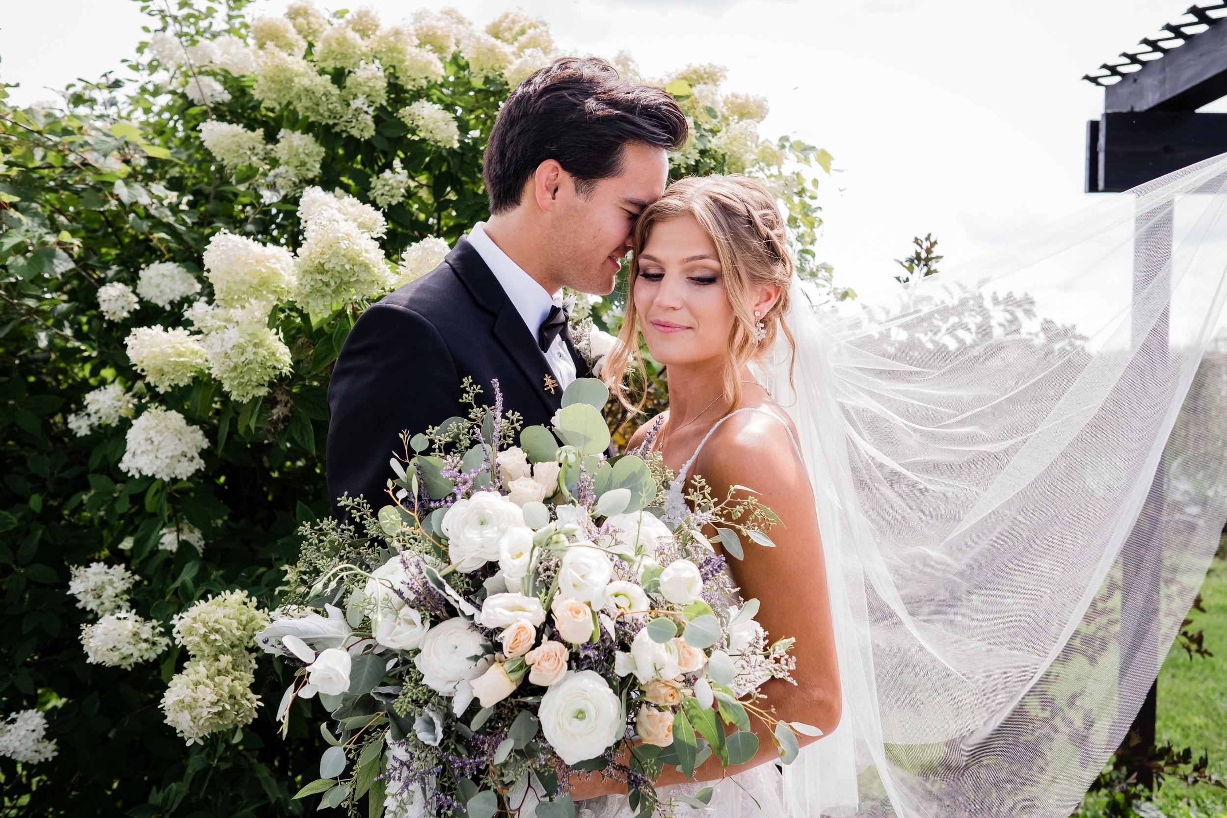 Newlywed couple standing close as the groom gently touches his forehead to the bride’s, who holds a large white and blush bouquet with greenery – her veil flowing in the breeze, surrounded by white flowering bushes at One Salon & Bridal Co