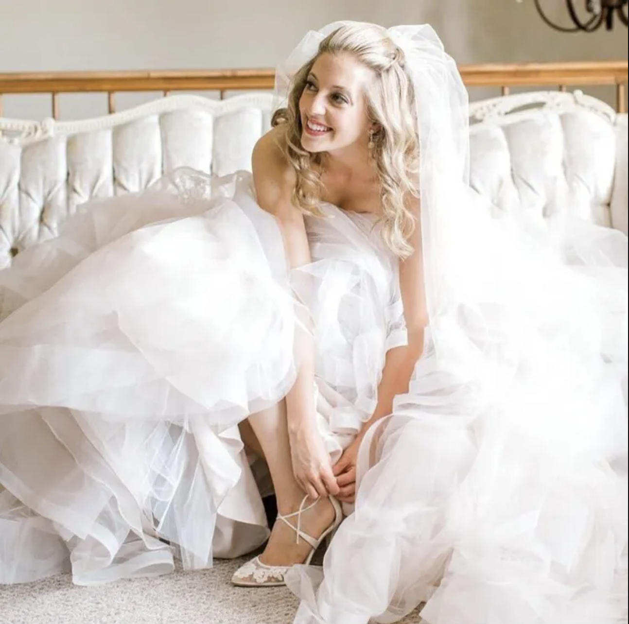 Bride in a wedding dress sitting on a bed, smiling as she adjusts her shoe – styled by One Salon & Bridal Co in St. Charles, IL