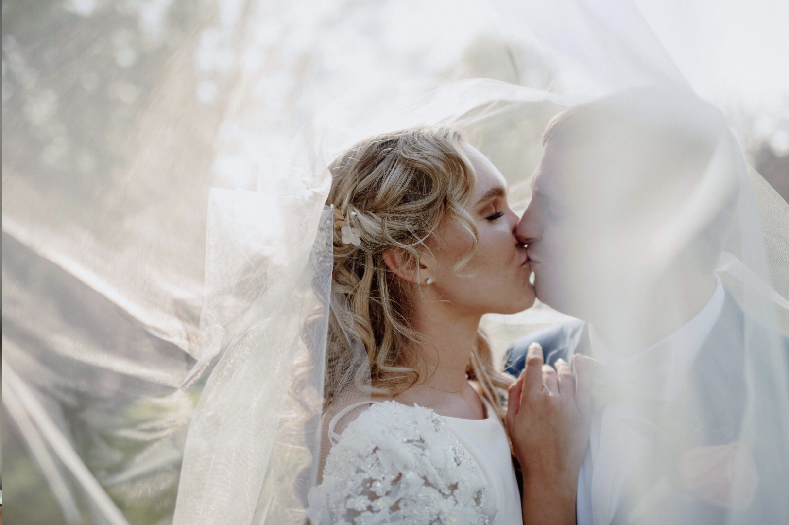 A bride and groom kiss under a sheer veil during their wedding, with natural outdoor light illuminating their faces--styled by One Salon & Bridal Co in St. Charles, IL