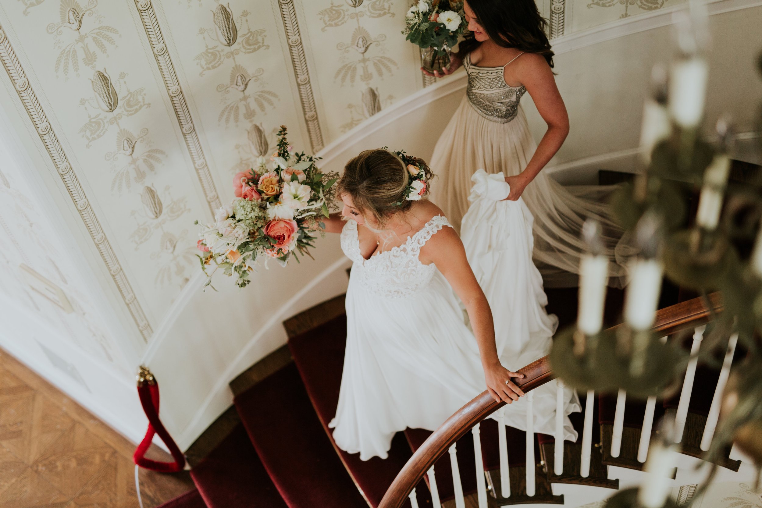 A bride in a white wedding dress holding a bouquet of pink, white, and green flowers descending a curved staircase with another woman in a beige dress – styled by One Salon & Bridal Co in St. Charles, IL