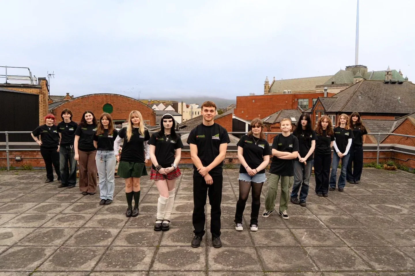 A group of seventeen teenagers and their mentor on a rooftop, all wearing black T-shirts with a green and white volume control graphic and text, smiling and standing in a semi-circle.