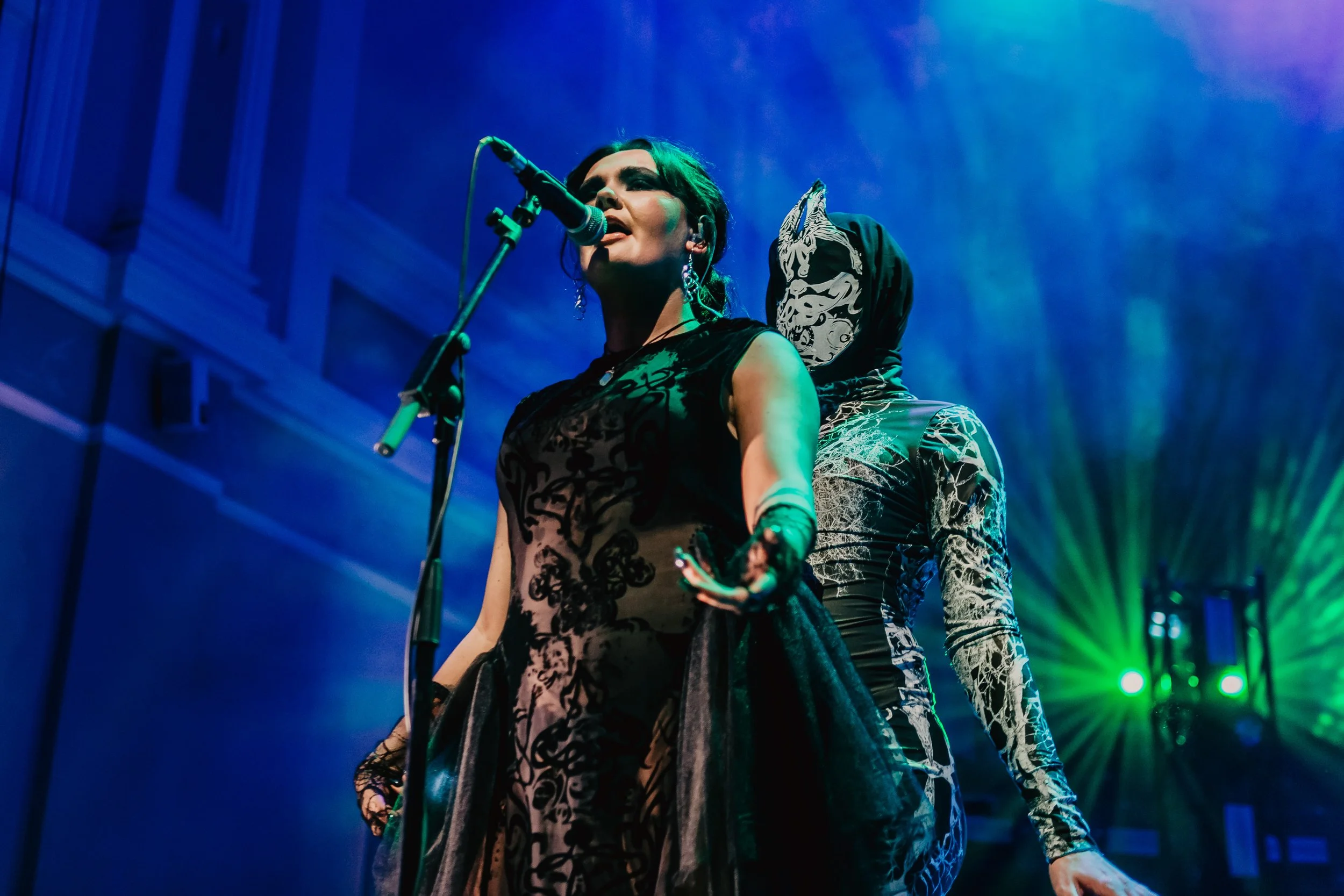 A women singing into a microphone in a dark lace dress with a masked figure looming behind her.