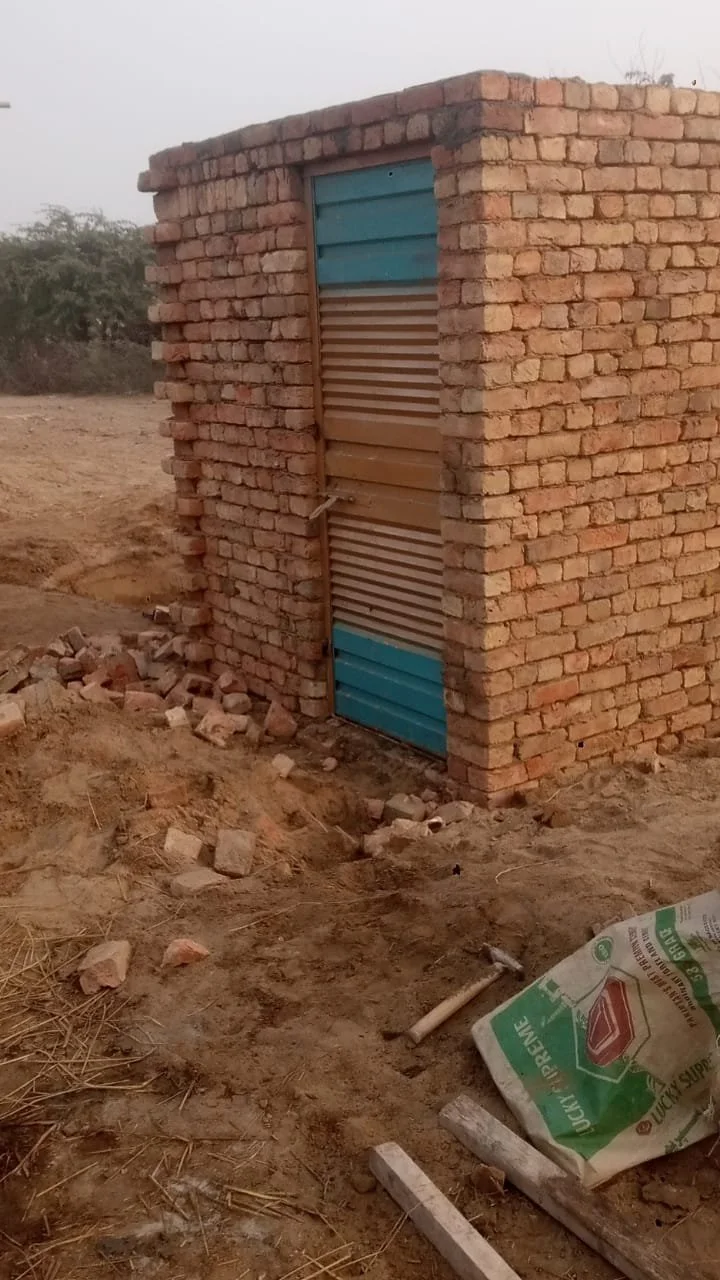 Small brick structure with a door made of colorful metal sheets in a rural area with dirt ground and construction debris.