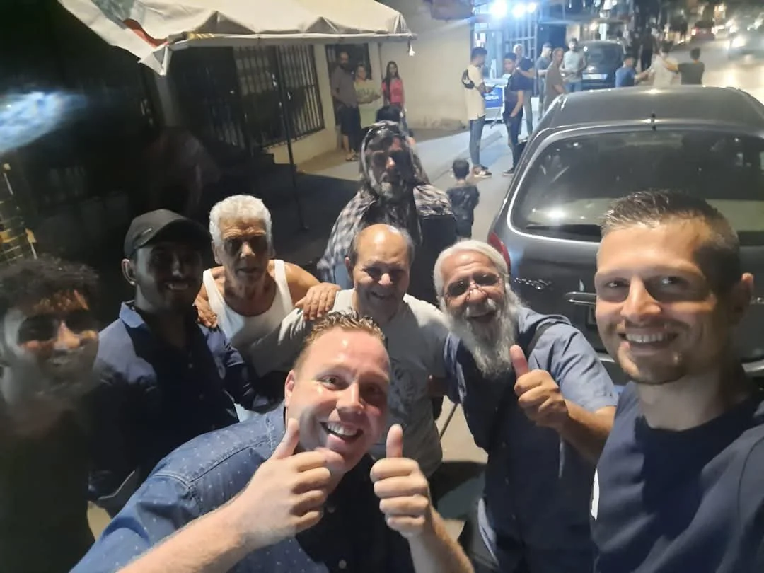 A group of smiling men taking a selfie on a city street at night, with cars and pedestrians in the background.