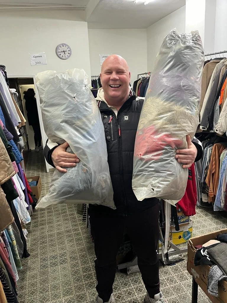 A man smiling and holding two large plastic bags filled with clothing in a thrift store aisle surrounded by racks of clothes.