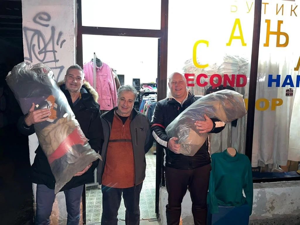 Three men standing inside a secondhand clothing store, each holding large plastic bags filled with clothes. The store has clothing racks and a mannequin with a green sweater.