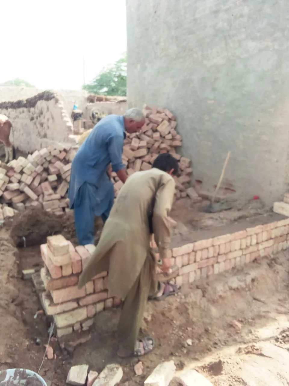 Two men building a brick wall outdoors, with piles of bricks nearby, in a construction site.