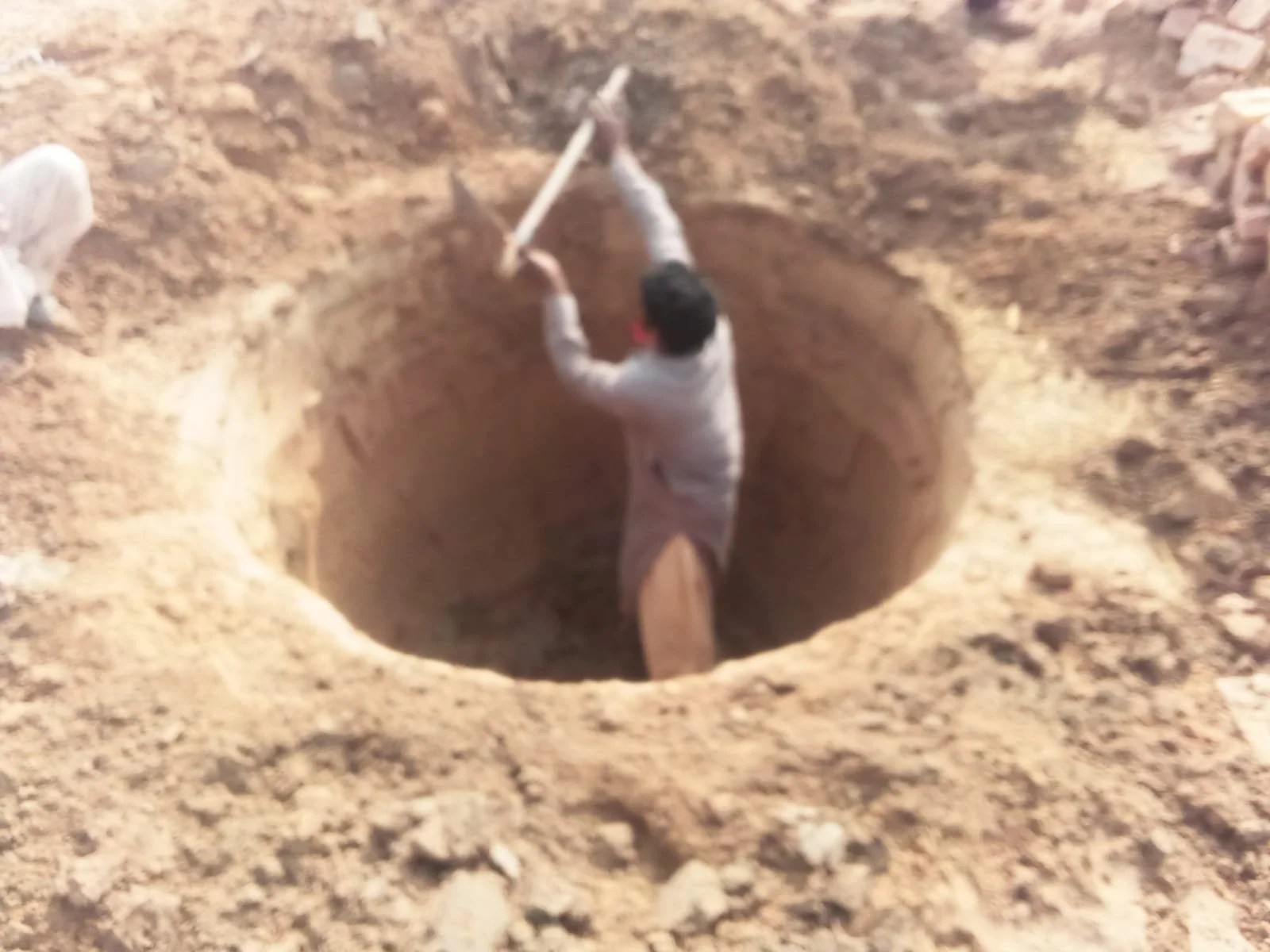 Man digging a hole in the ground with a shovel, surrounded by loose dirt and rocks.